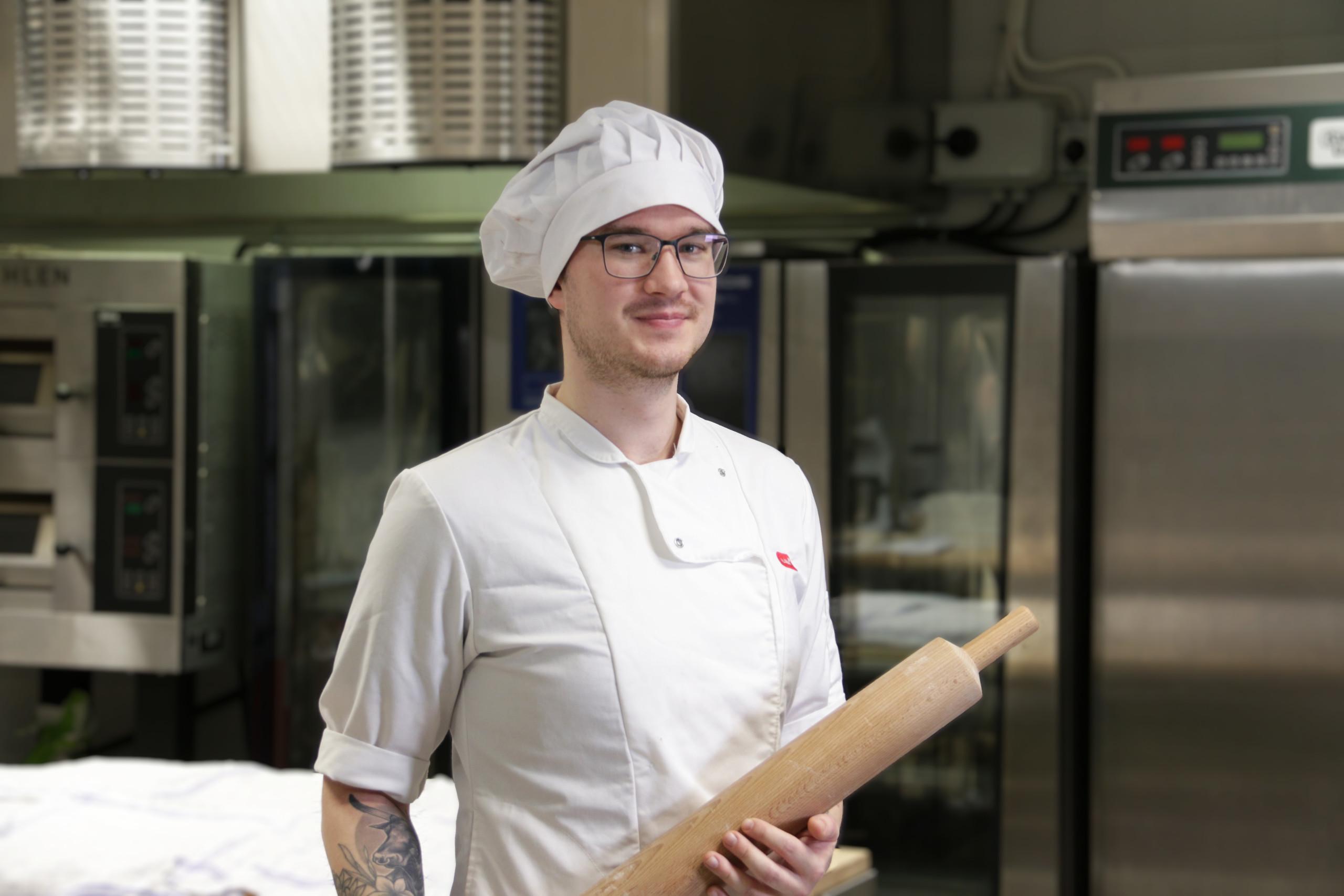 Leipuri-kondiittoriopiskelija valkoisessa leipuriasussa kaulin kädessään.