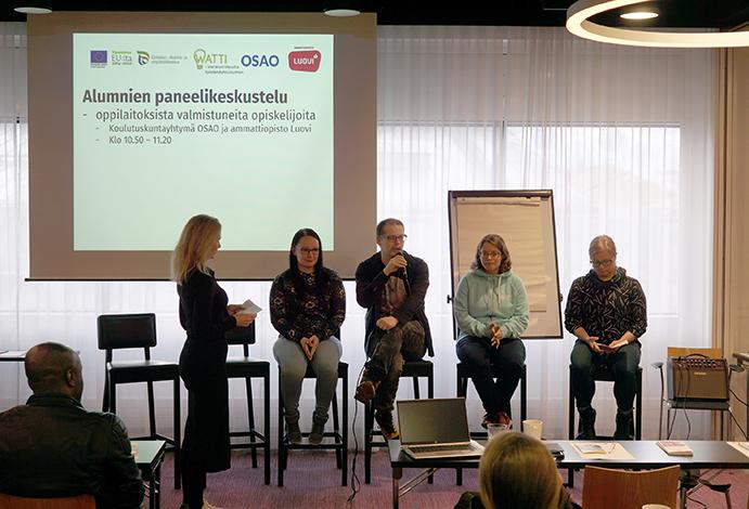 Neläj henkilöä istuu huoneen etualalla, vieressä seisoo juontaja Sari Kauppila. Takana näkyy valkokankaalla esitys, jossa lukee "Alumnien paneelikeskustelu".