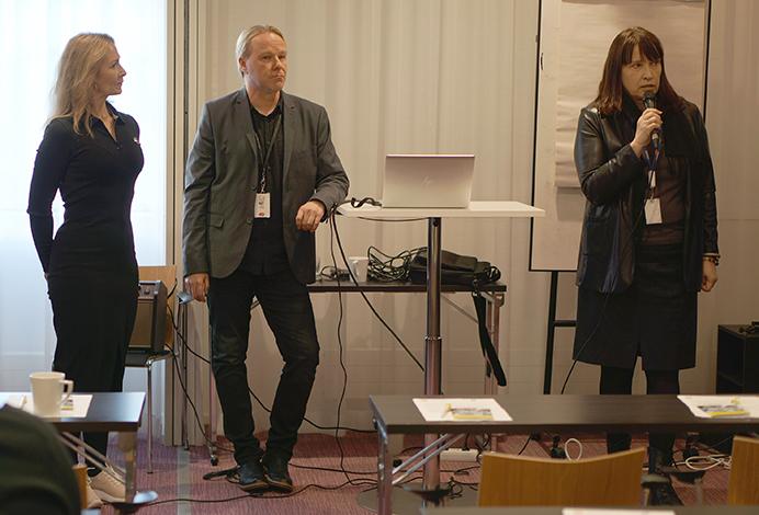 Tilaisuuden juontaja Sari Kauppila, hankepäällikkö Marko Ylisirkka ja hankekehityspäällikkö Merja Paloniemi seisovat huoneen edessä, Merja Paloniemi puhuu mikrofoniin.