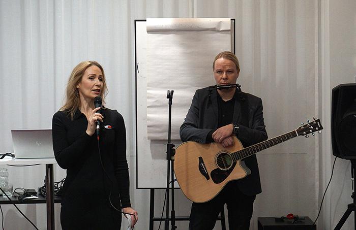 Tilaisuuden juontaja Sari Kauppila puhuu mikrofoniin ja WATTI-hankkeen hankepäällikkö Marko Ylisirkka kitara sylissään.