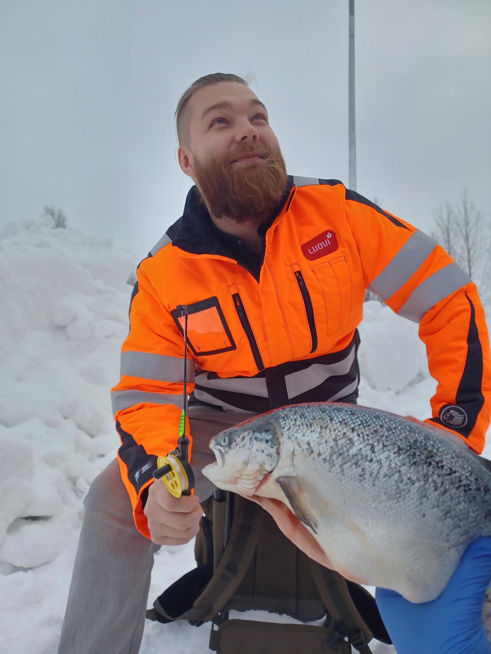 Luovin työntekijä istuu ylpeänä pilkkivapa kädessään saaliina perattu lohi.