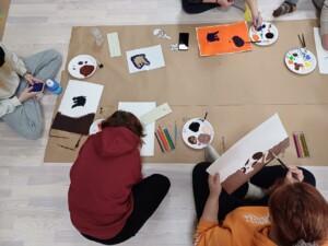Henkilöitä istuu lattialla paperien ja maalien ääressä maalaten.