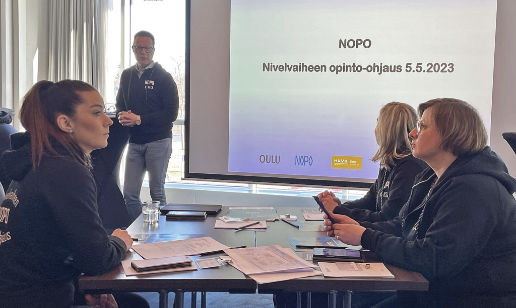 Projektipäällikkö Timo Kettunen seisoo valkokankaan vieressä, jossa lukee NOPO Nivelvaiheen ohjaus 5.5.2023. Edessä pöytä, jonka ääressä istuu 3 ihmistä.