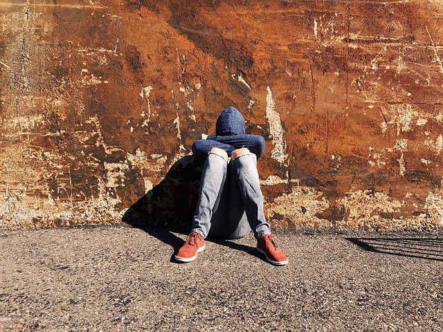 Nuori henkilö istuu yksin selkä seinää vasten, pää hupun sisällä ja pää alas painuneena.