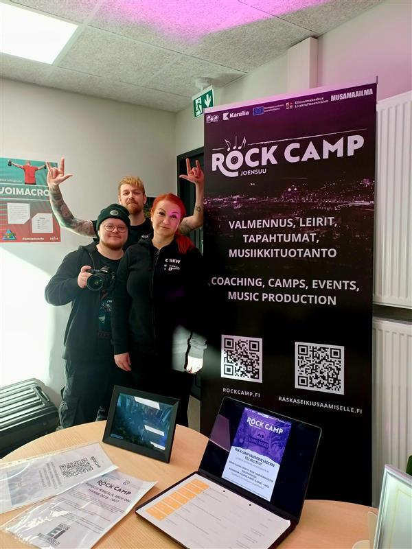Kolme ihmistä mustissa vaatteissa katsoo kameraan, vieressä roll up, jossa lukee "Rock Camp Joensuu - valmennus, leirit, tapahtumat, musiikkituotanto - coaching, camps, events, music production".
