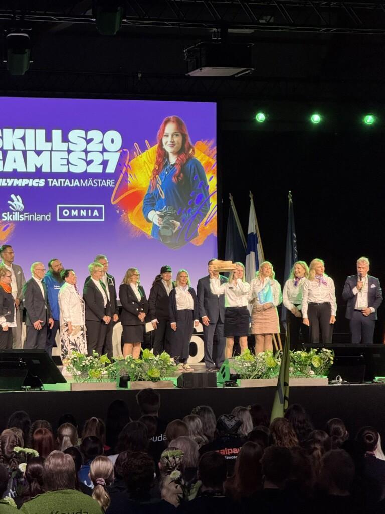 Lavalla paljon ihmisiä, Luovin rehtori Seija Norup nostaa Taitaja-kapulaa. Takana lukee Skills Games 27.