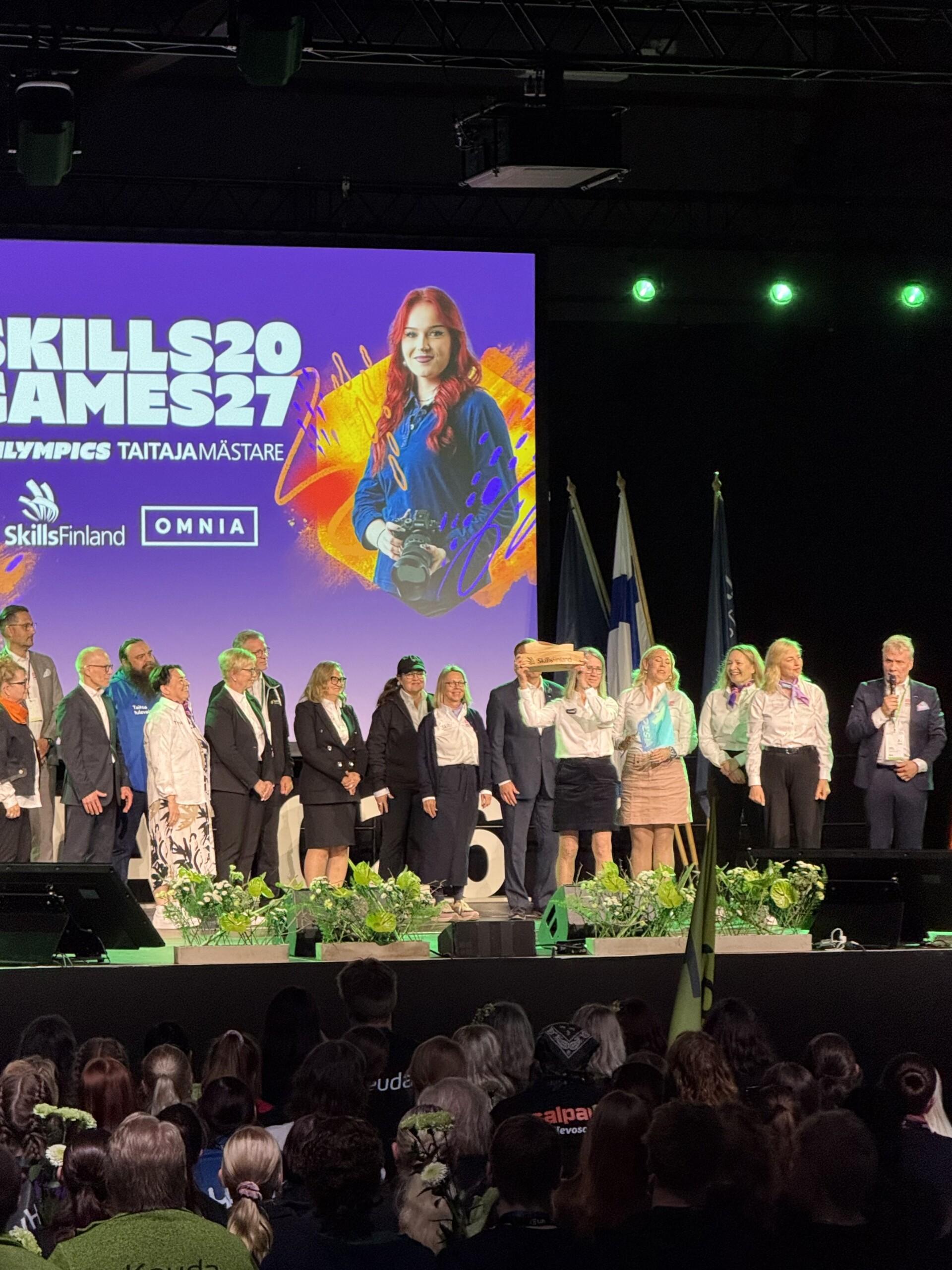 Lavalla paljon ihmisiä, Luovin rehtori Seija Norup nostaa Taitaja-kapulaa. Takana lukee Skills Games 27.