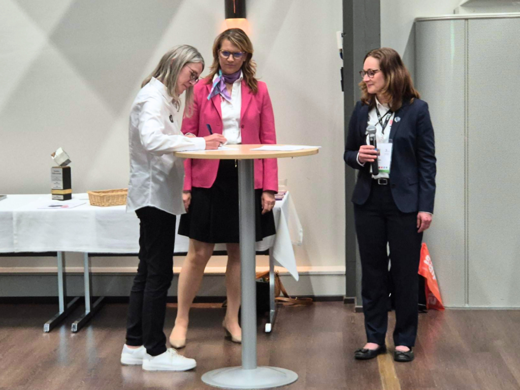 Seija Norup allekirjoittaa paperia korkean pöydän ääressä, takana Maria Ekroth ja Katri Pokkinen.