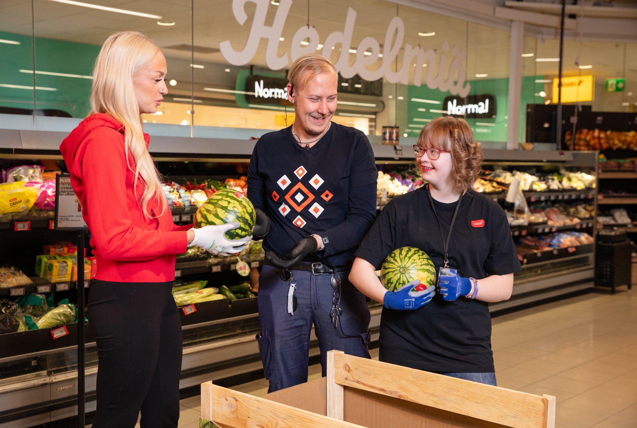 Luovin ohjaaja punaisessa Luovi-hupparissaan, päivittäistavarakaupan työpaikkaohjaaja työasussaan ja Luovin opiskelija työasussaan päivittäistavarakaupassa hyllyttävät vesimeloneja.