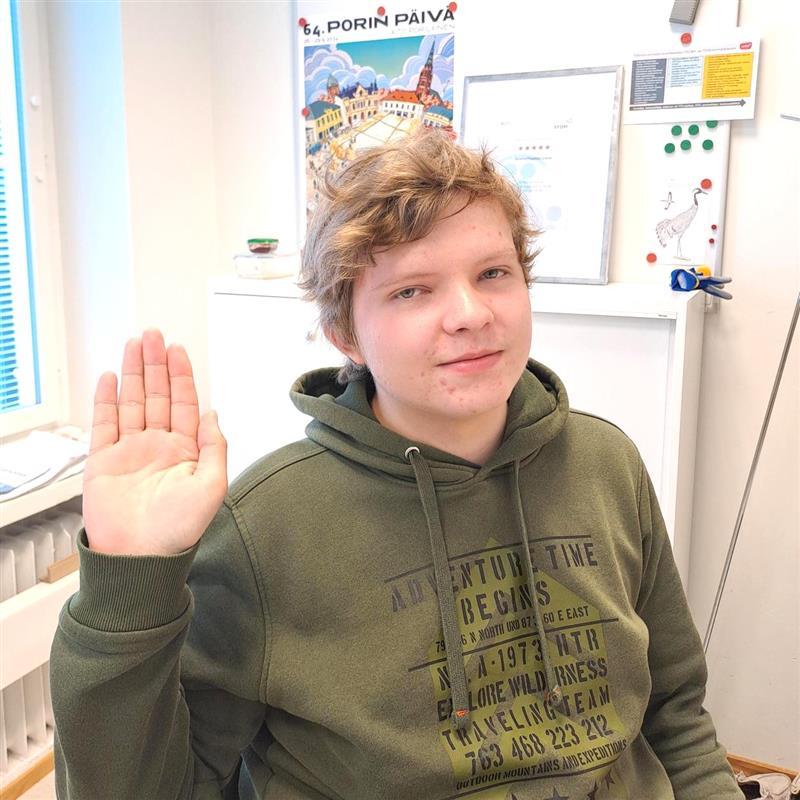 Nuori henkilö hymyilee kameralle ja nostaa kättään.
