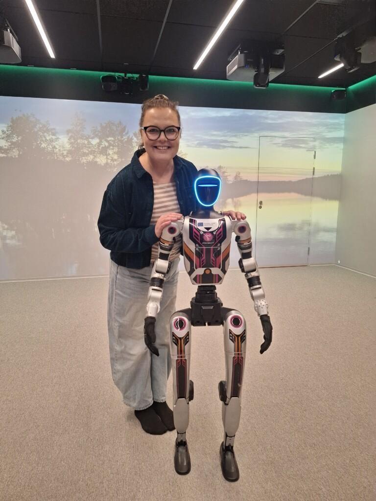 Piia Jokelainen katsoo hymyillen kameraan ja pitelee hartioista kahdella jalalla seisovaa robottia.