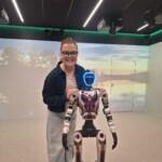 Piia Jokelainen katsoo hymyillen kameraan ja pitelee hartioista kahdella jalalla seisovaa robottia.