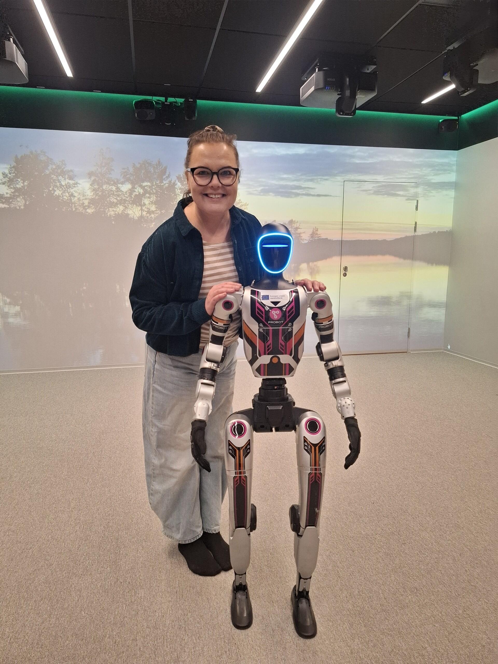 Piia Jokelainen katsoo hymyillen kameraan ja pitelee hartioista kahdella jalalla seisovaa robottia.