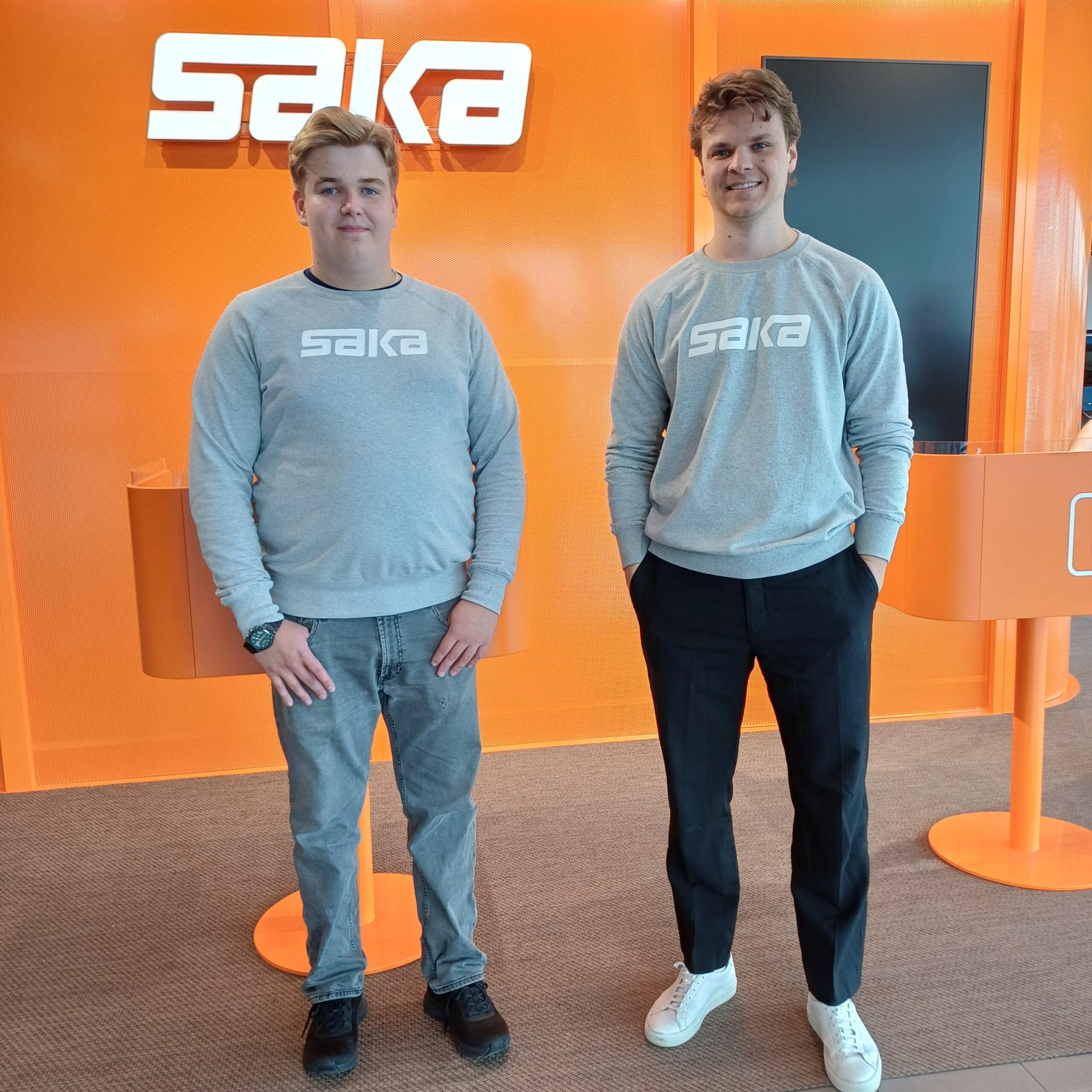 Alex ja Rasmus Nordlund autoliike Sakan tiloissa, päällää harmaat Saka-logolliset paidat ja takana oranssi seinä, jossa Saka-logo.