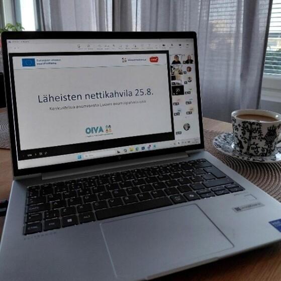Pöydällä kannettava tietokone ja kahvikuppi, näytöllä näkyy Teams-kokous, jonka esityksessä lukee "Läheisten nettikahvila 25.8.".