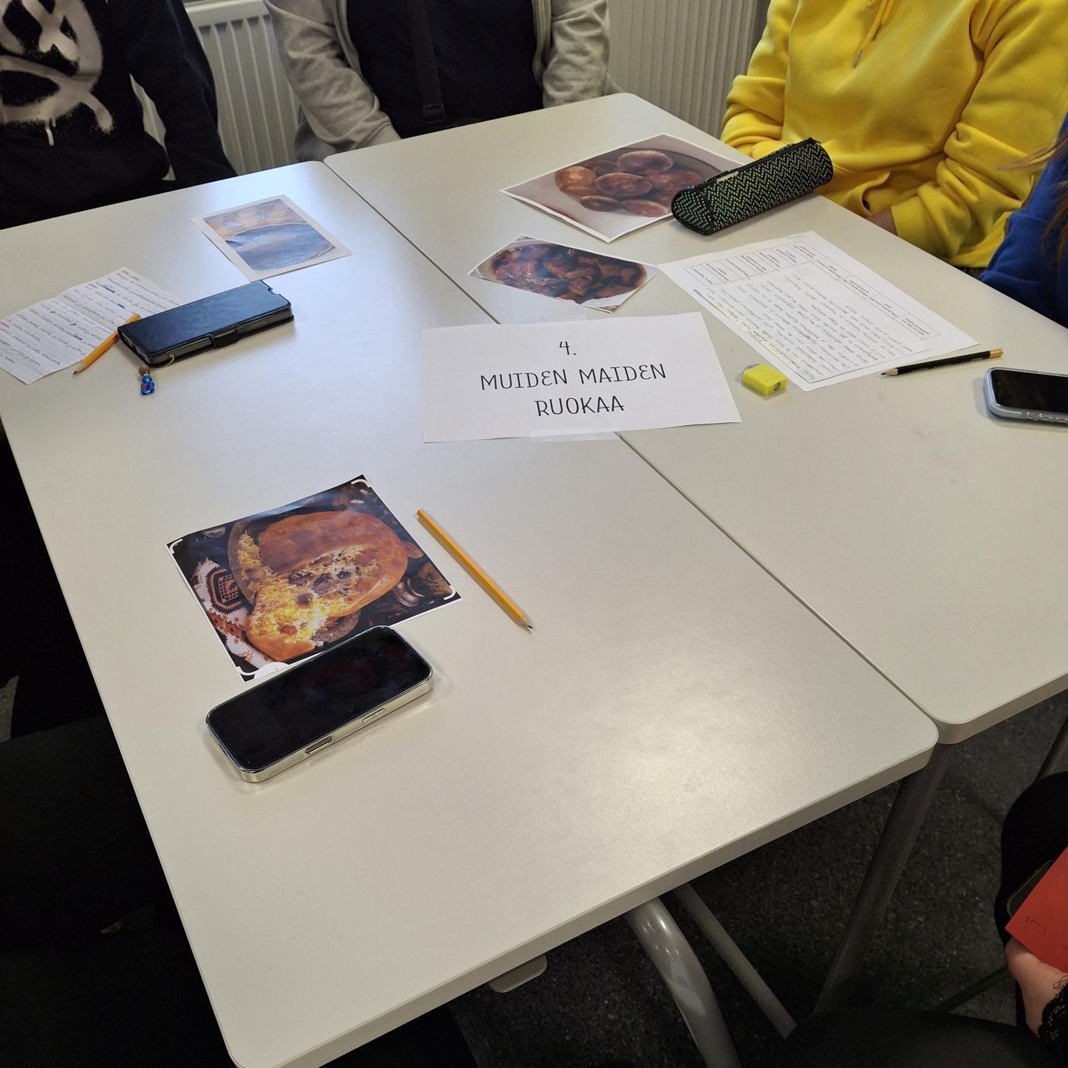 Pöydällä kuvia ruoista sekä paperi, jossa teksti "Muiden maiden ruokia", pöydän ääressä istuu ihmisiä.
