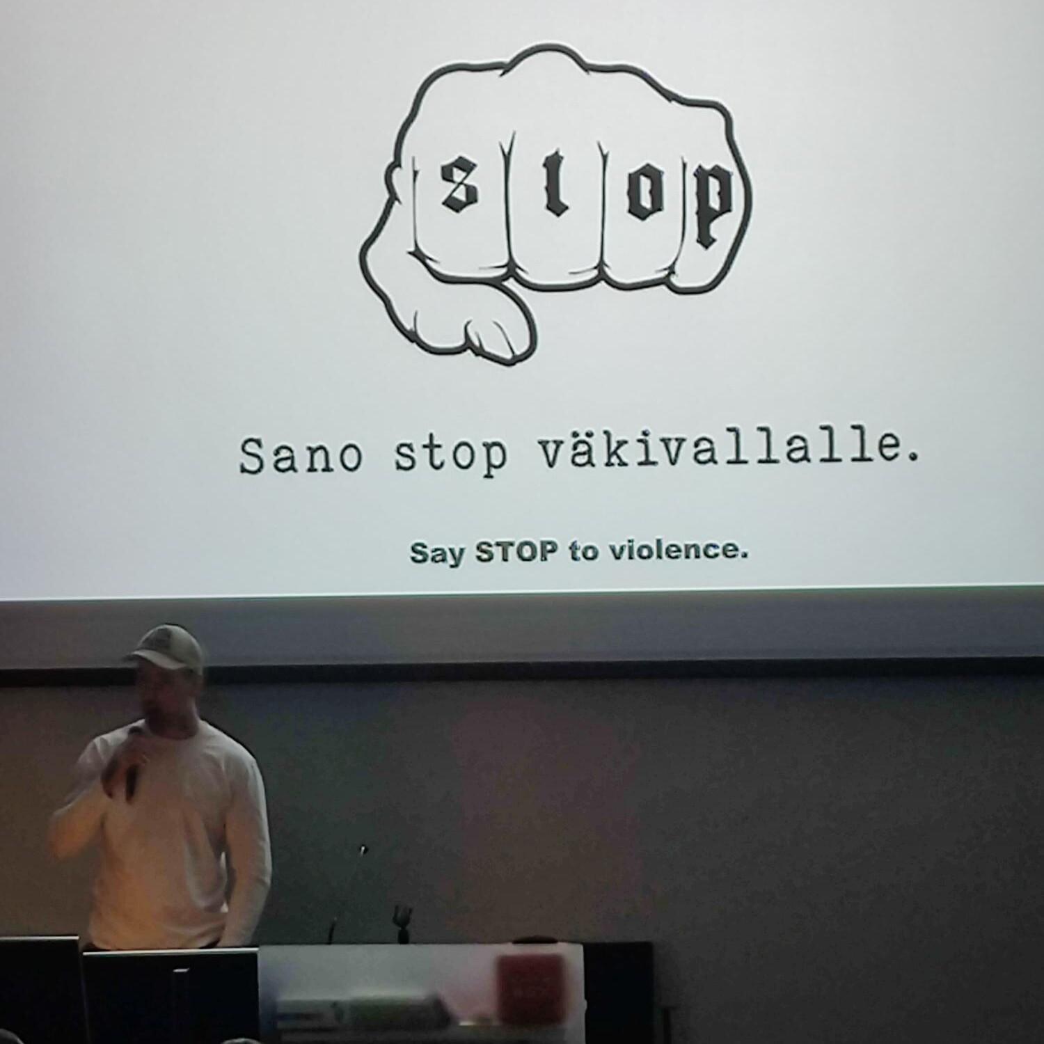 Henkilö puhuu yleisölle, takana esitys jossa lukee "Sano stop väkivallalle" ja näkyy nyrkki, jonka rystysissä lukee "stop".